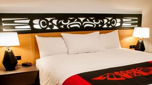 Una habitación de hotel con una cama grande, ropa de cama blanca, una manta roja y negra, dos lámparas de noche y un cabecero moderno.