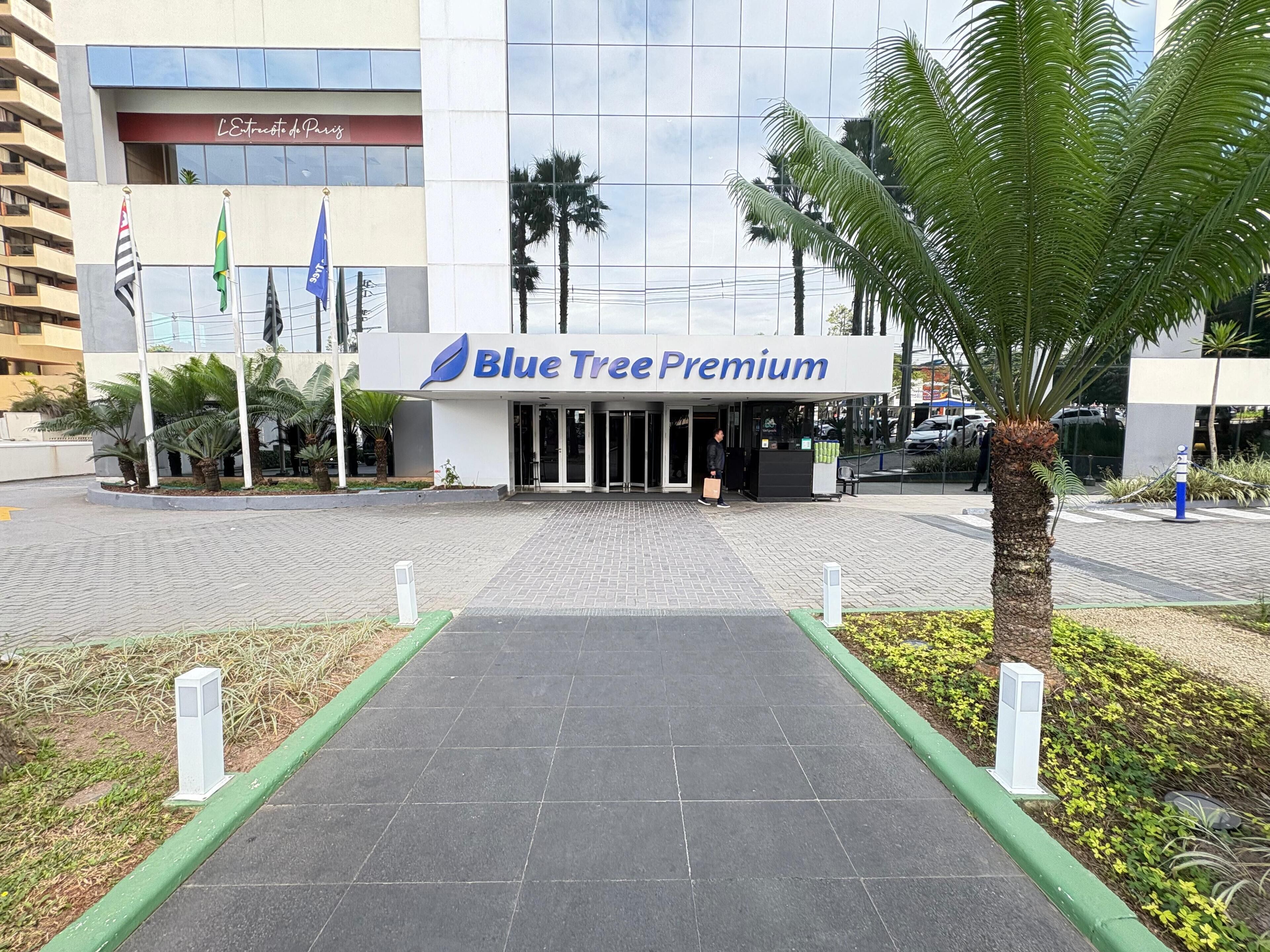 The entrance to Blue Tree Premium with a paved pathway leading to the entrance, palm trees, and flags.