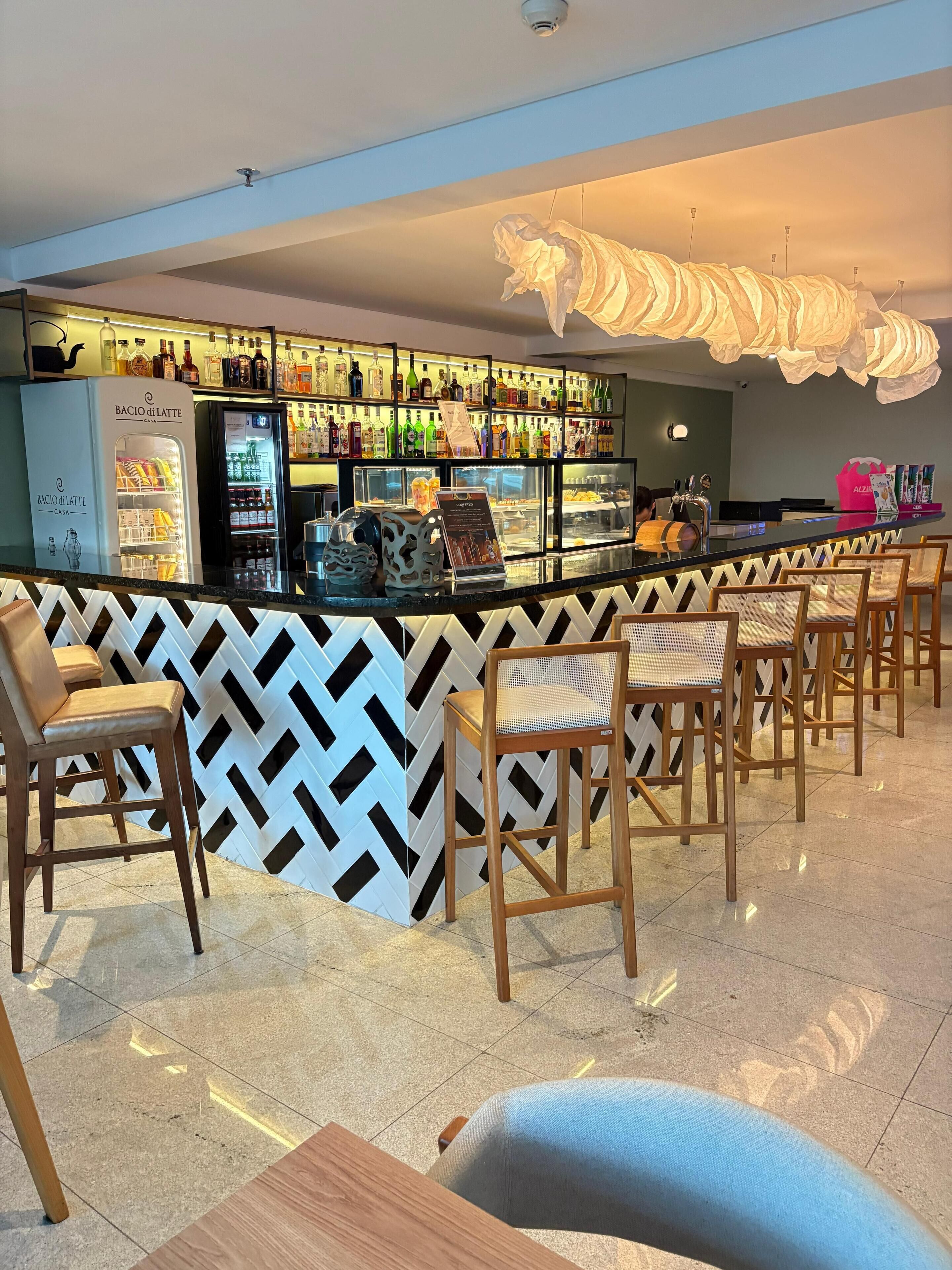 Uma área de bar com parede em chevron preta e branca, uma geladeira e vários frascos. Há cadeiras de madeira e um balcão com uma pessoa atrás.