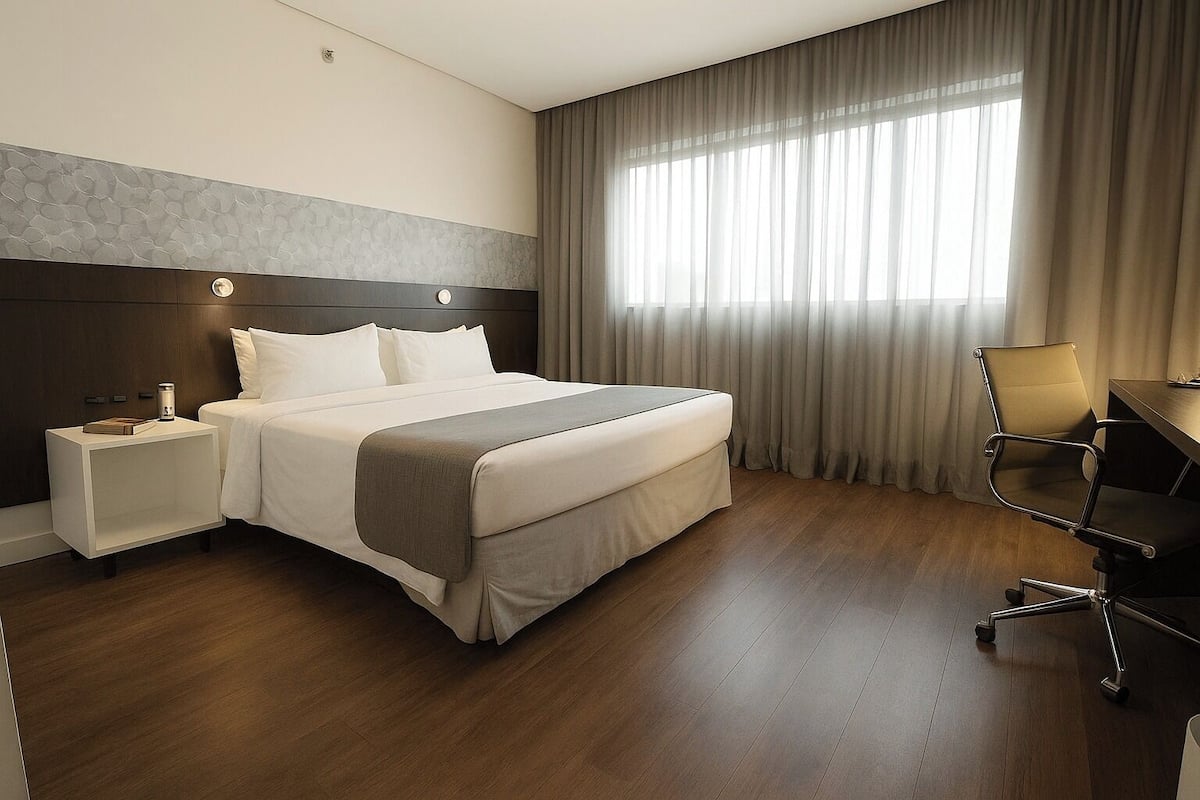 A hotel room with a large bed, a nightstand, a desk with a chair, and a window with curtains.