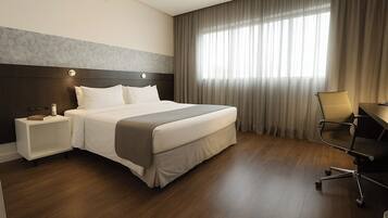 A hotel room with a large bed, a nightstand, a desk with a chair, and a window with curtains.