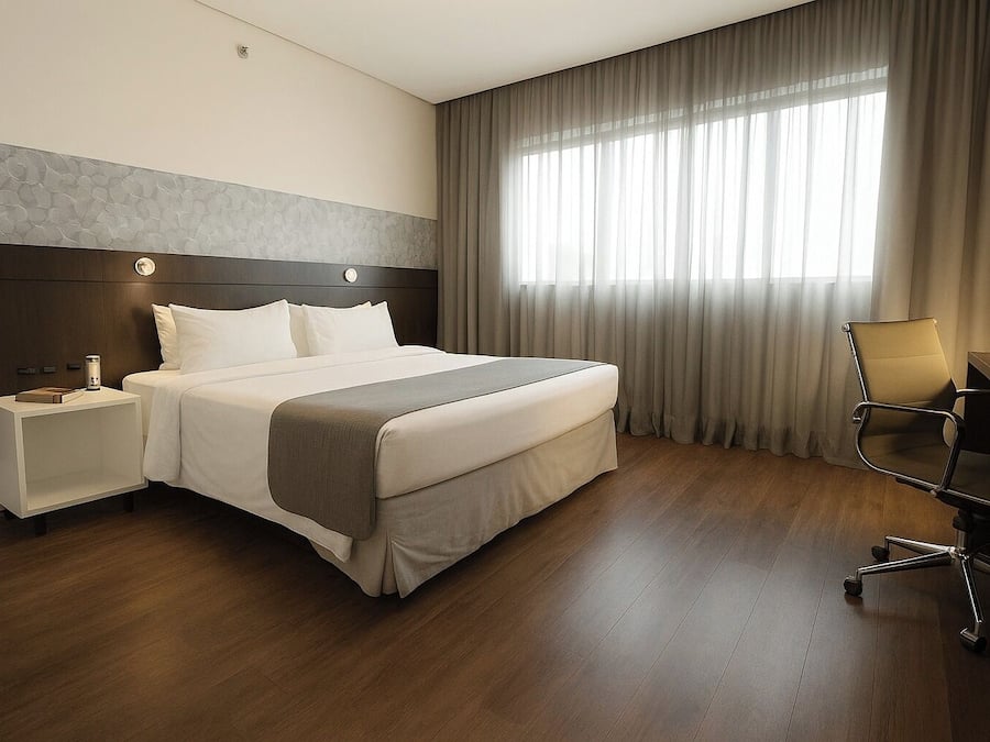A hotel room with a large bed, a nightstand, a desk with a chair, and a window with curtains.