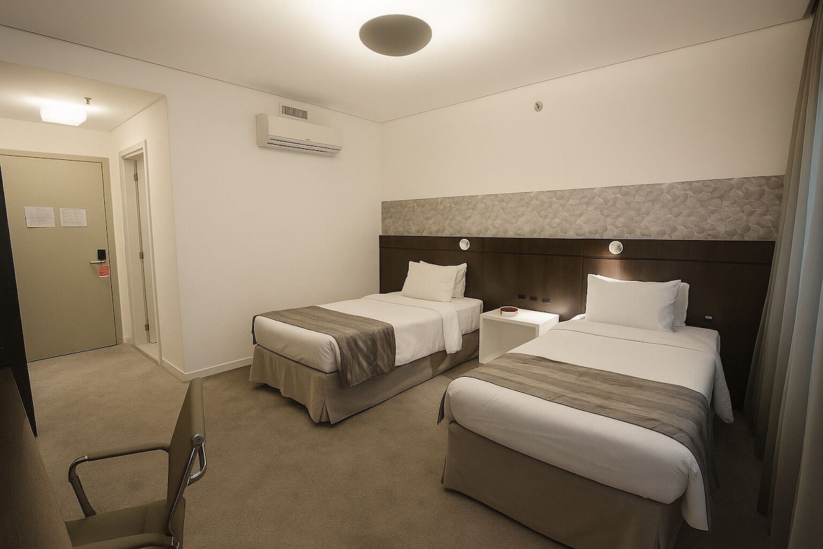 A hotel room with two beds, a bedside table, an air conditioning unit, a door, and a chair.