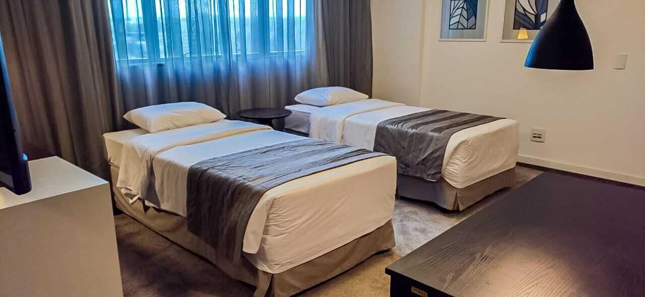 Quarto de hotel com duas camas, uma mesinha e uma janela grande com cortinas.