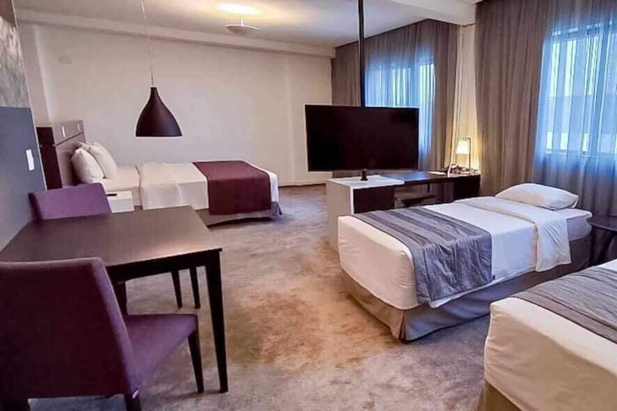 A hotel room with two beds, a desk, a TV, and a large window with curtains.