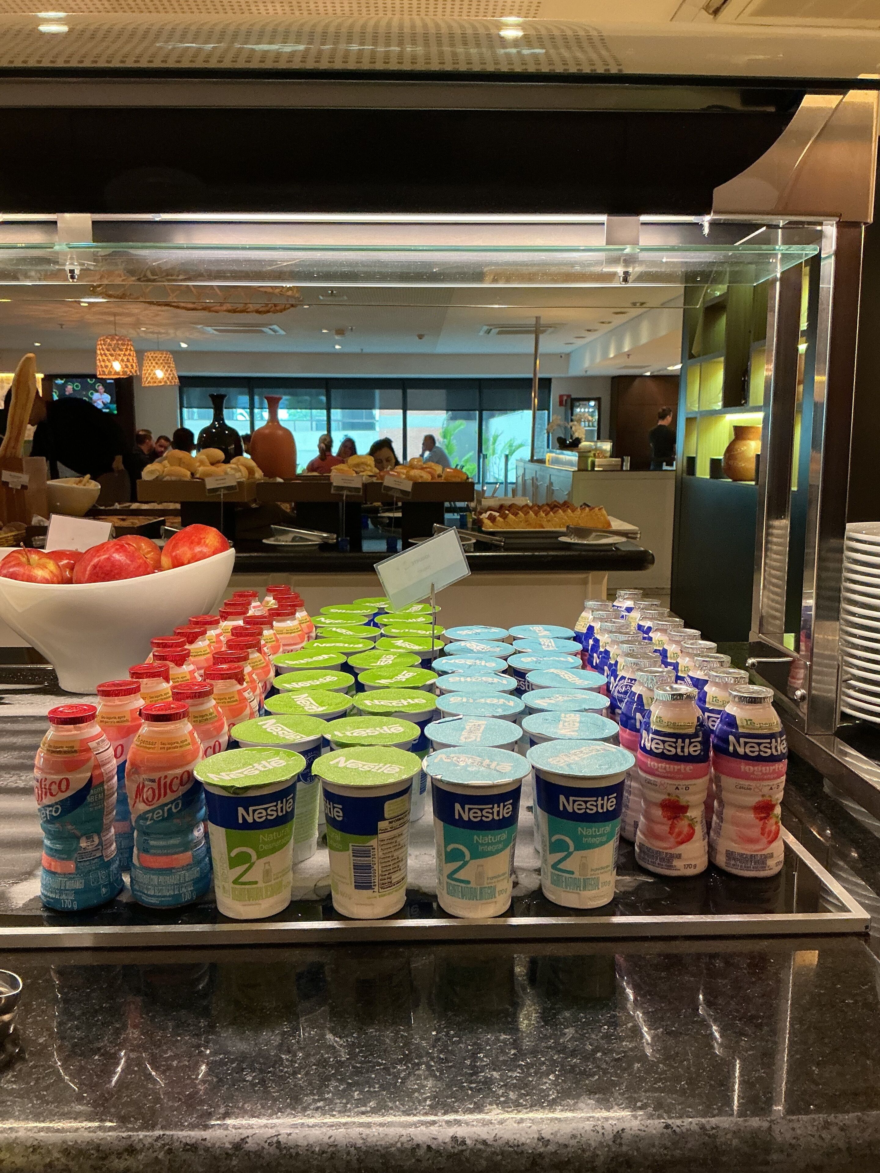 Uma área de buffet com diversas opções de comida e bebida, incluindo uma tigela de frutas, garrafas de água e potes de iogurte Nestlé.