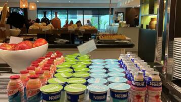 Uma área de buffet com diversas opções de comida e bebida, incluindo uma tigela de frutas, garrafas de água e potes de iogurte Nestlé.