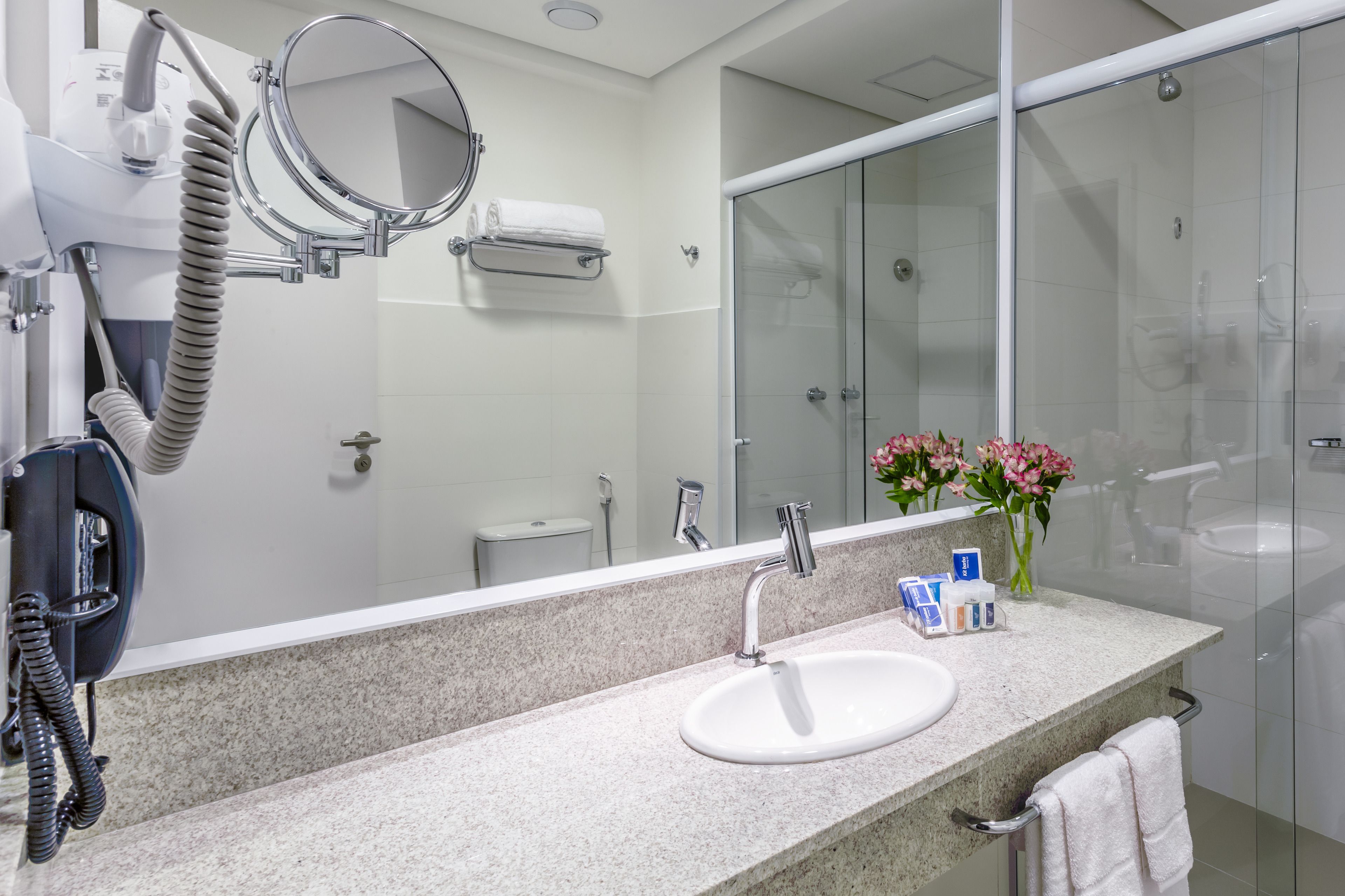 Une salle de bain moderne avec un comptoir en granit, un lavabo blanc, un sèche-cheveux portatif, un miroir rond et une cabine de douche en verre.