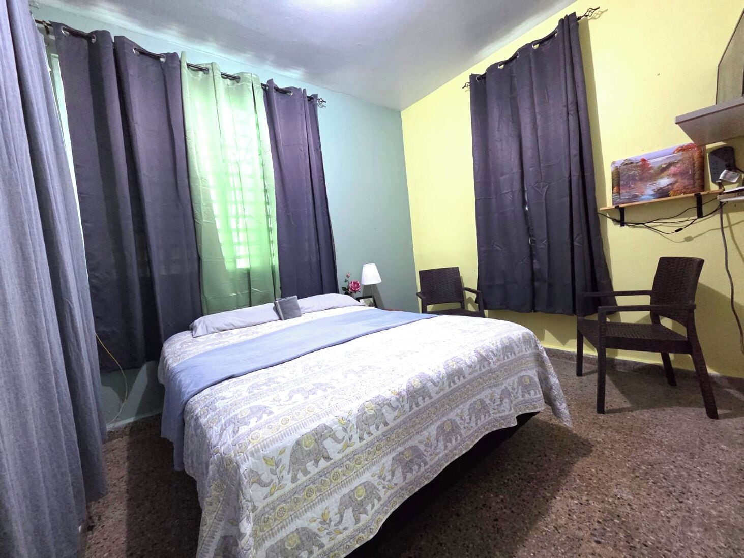 A hotel room with a bed, two chairs, a desk with a lamp, and a TV on the wall.