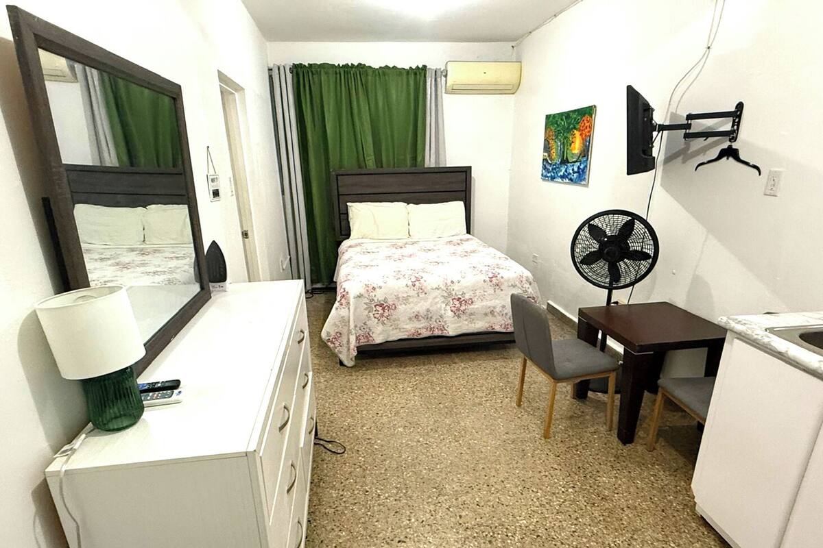 Ett hotellrum med en säng, ett skrivbord, en stol, en byrå, en lampa, en fläkt, en luftkonditioneringsanläggning och en tavla på väggen.