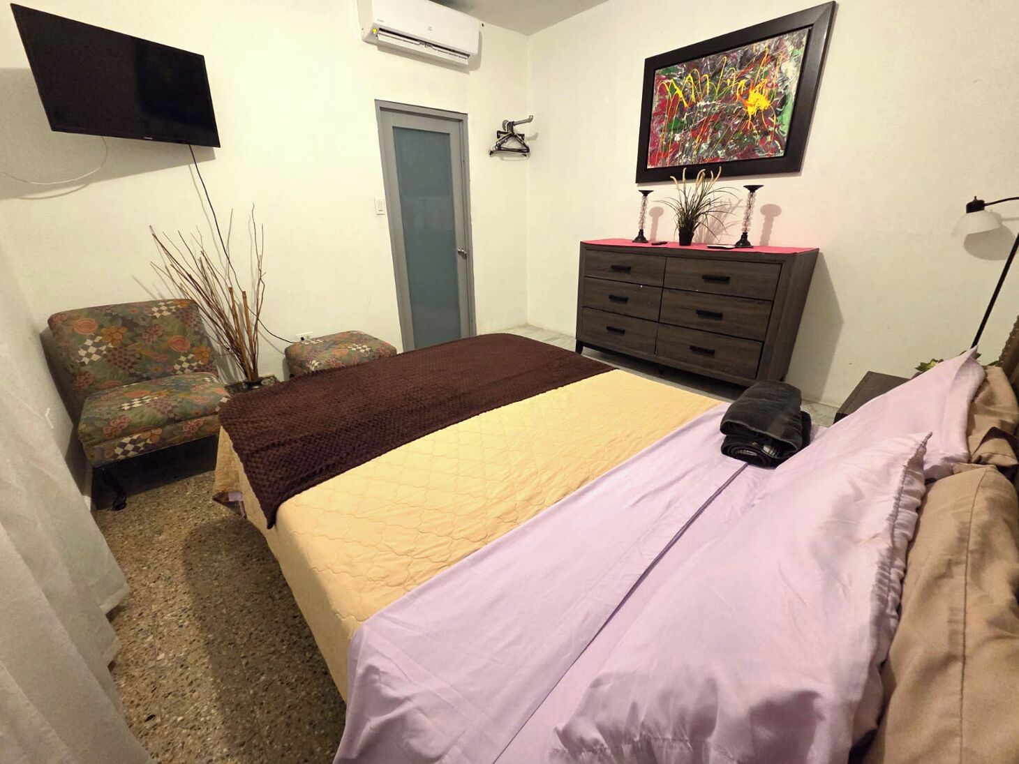 A bedroom with a bed, a chair, a dresser, a TV, and a door.