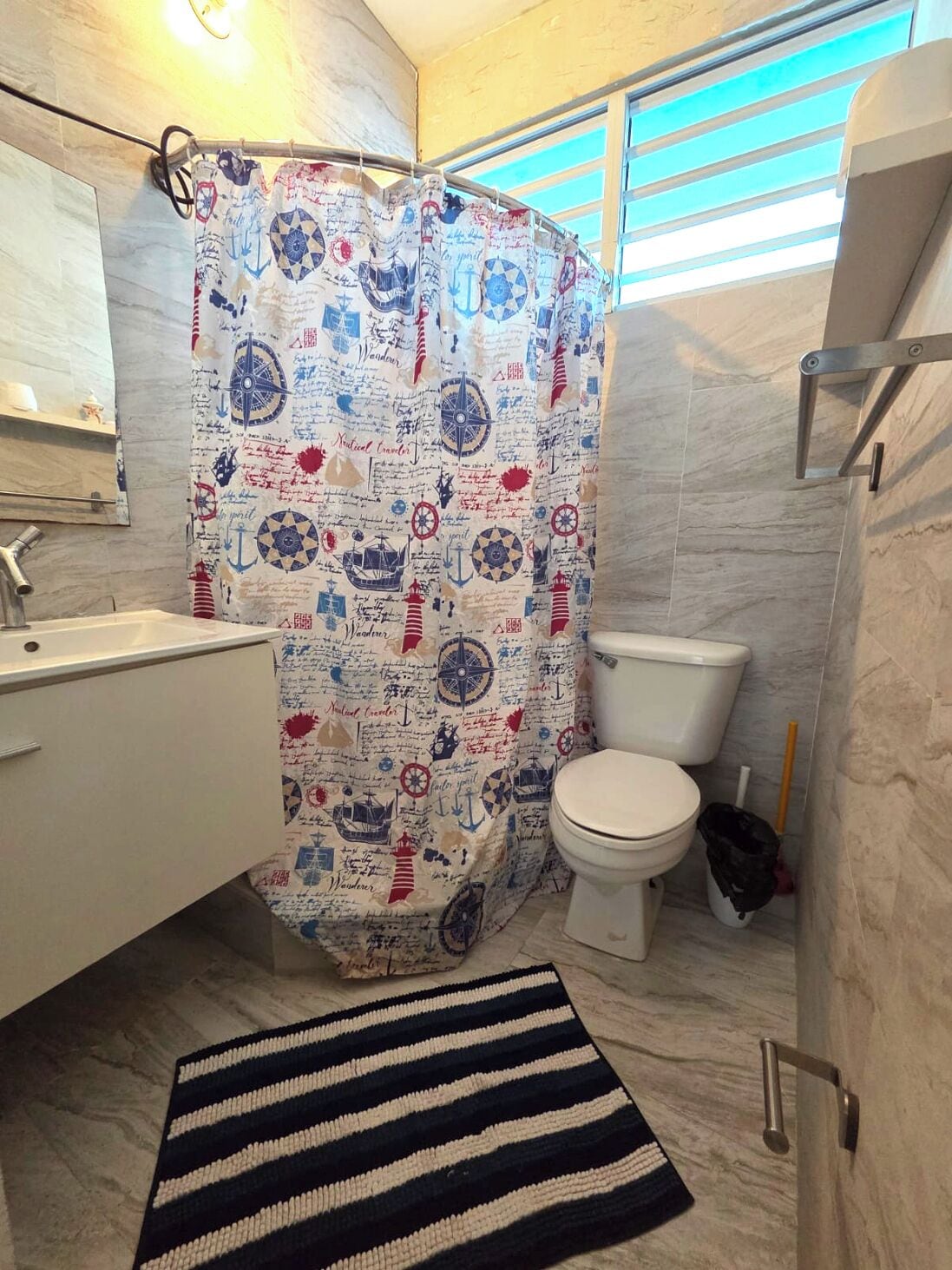 Een badkamer met een douchegordijn met nautisch thema, een wit toilet, een wastafel en een gestreept vloerkleed.