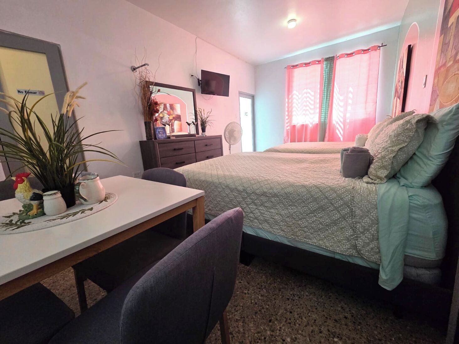 A small, cozy room with a bed, a desk, and a chair. There is a dresser with a mirror and a television mounted on the wall.