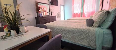 A small, cozy room with a bed, a desk, and a chair. There is a dresser with a mirror and a television mounted on the wall.