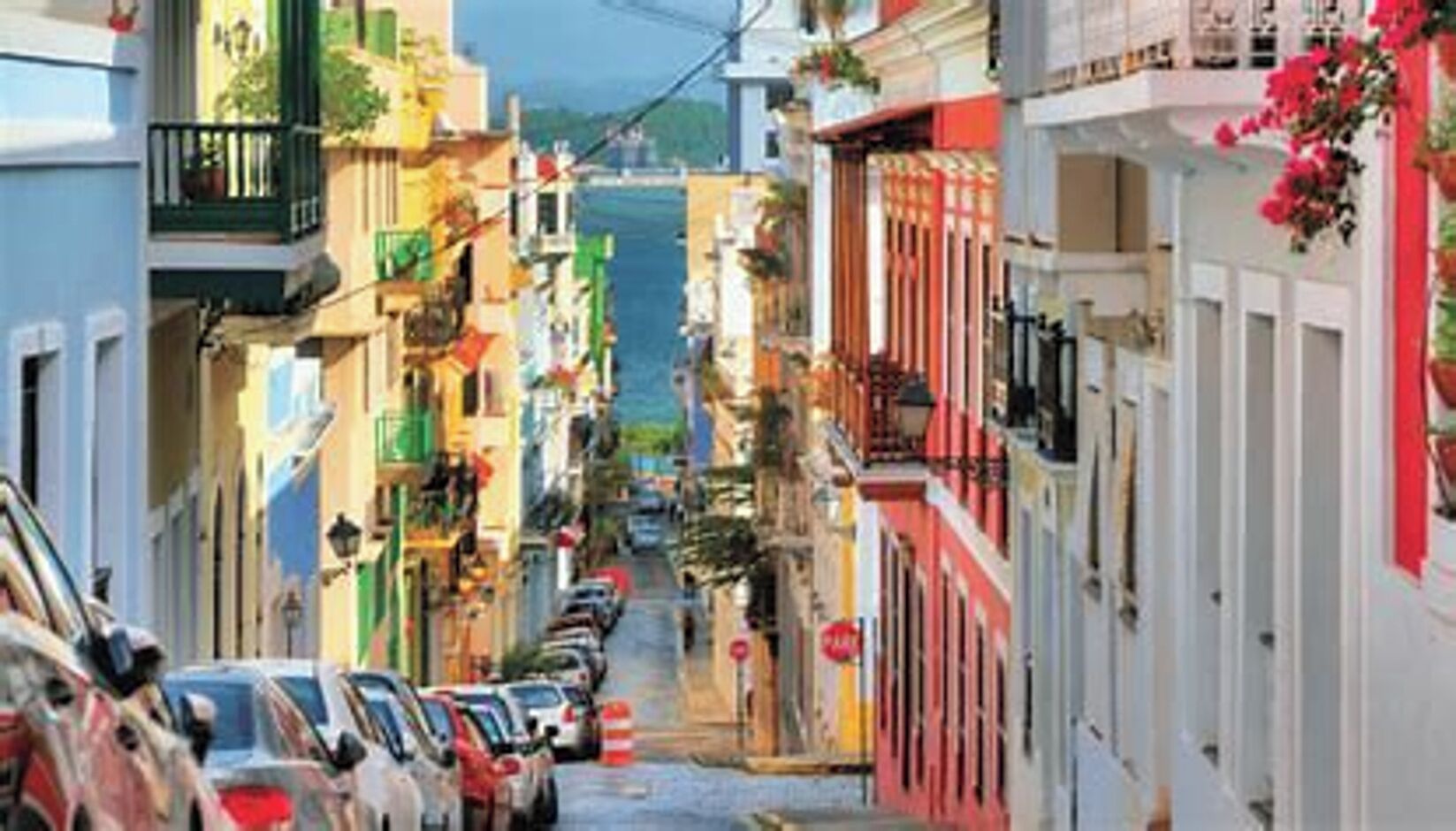 Une rue étroite bordée de bâtiments aux couleurs vives, de voitures garées et de balcons ornés de plantes.