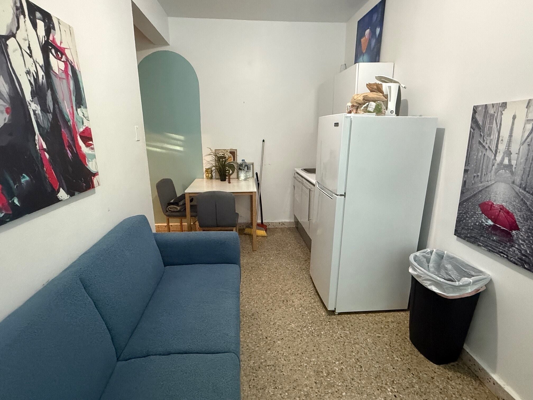 A compact kitchen with a refrigerator, a dining table with chairs, a blue sofa, and a trash bin.