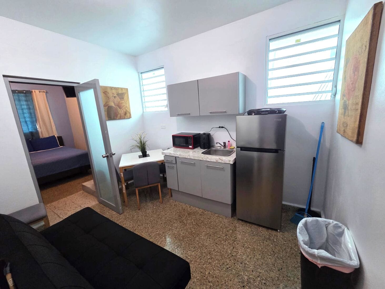 Cocina compacta con microondas, refrigerador y lavamanos. Hay una pequeña mesa de comedor con sillas, una planta en maceta y un cuadro en la pared.