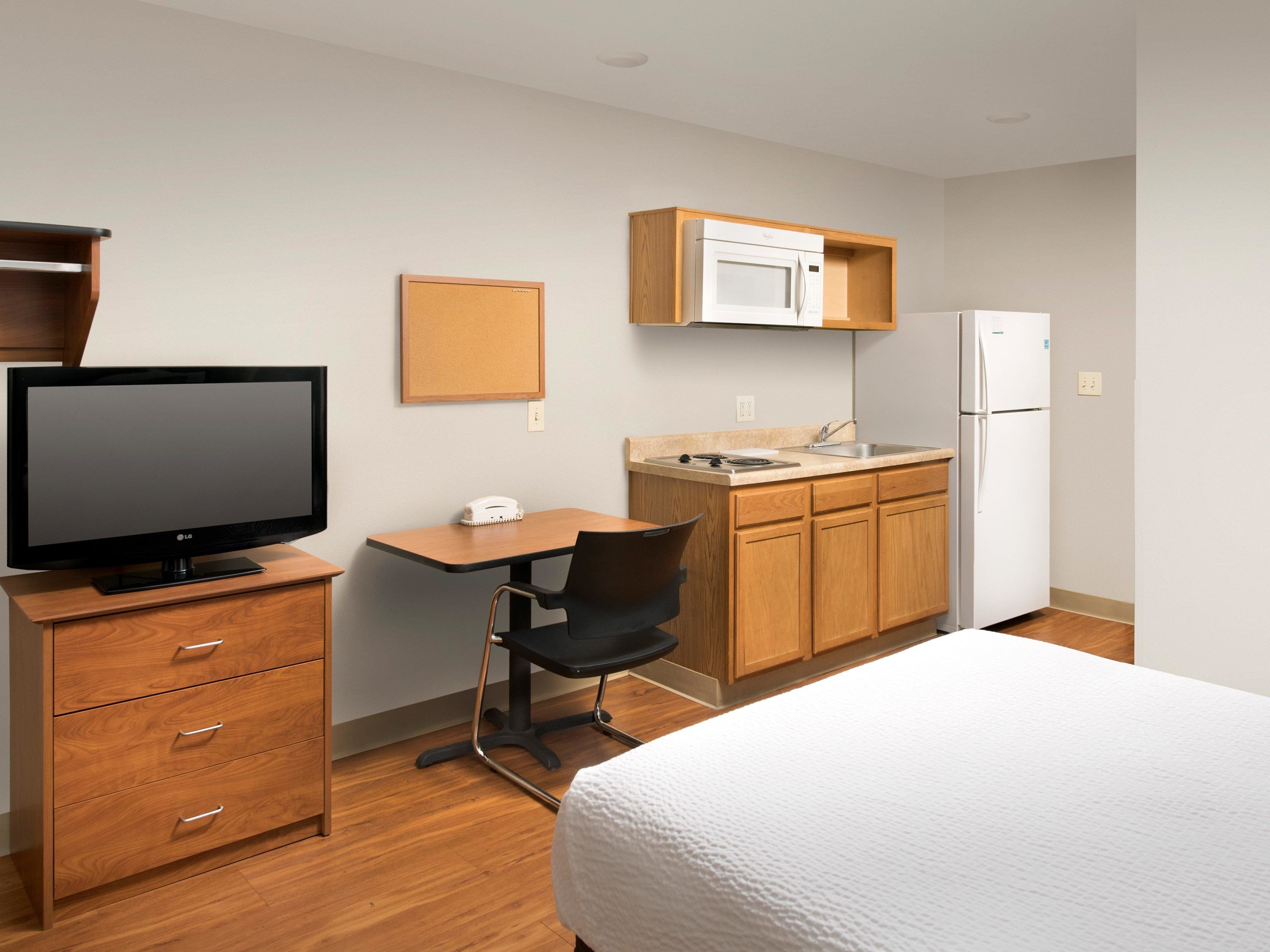 A hotel room with a bed, a desk with a chair, a television, a microwave, a refrigerator, and wooden cabinets.