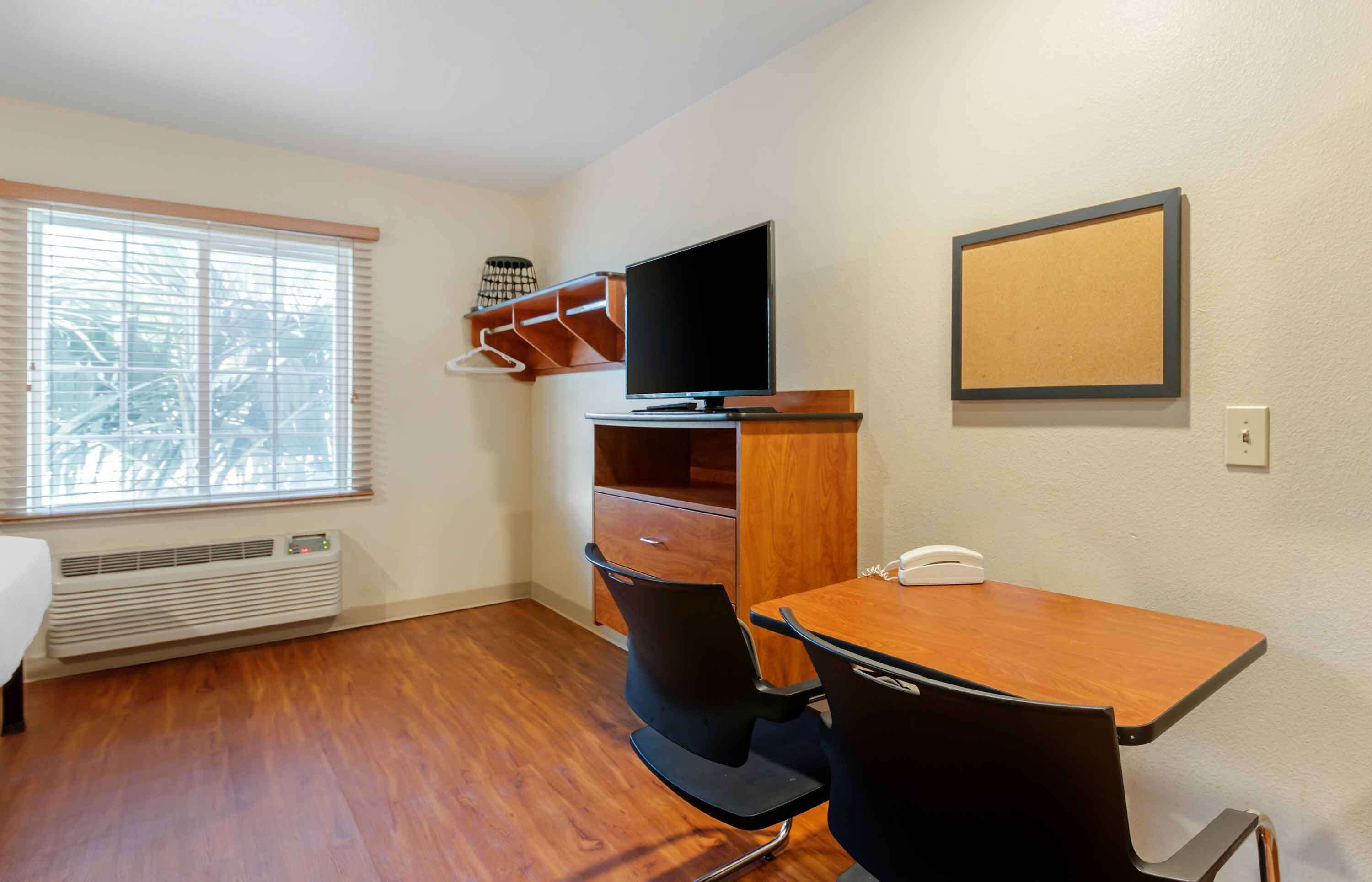 Une petite pièce meublée, avec un bureau en bois, deux chaises noires, un téléviseur posé sur un meuble en bois, un porte-manteau et une fenêtre avec des stores.
