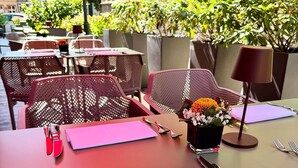 Ein Tisch ist gedeckt mit rotem Besteck, rosa Servietten und einer Blumenvase, umgeben von Stühlen und Topfpflanzen.