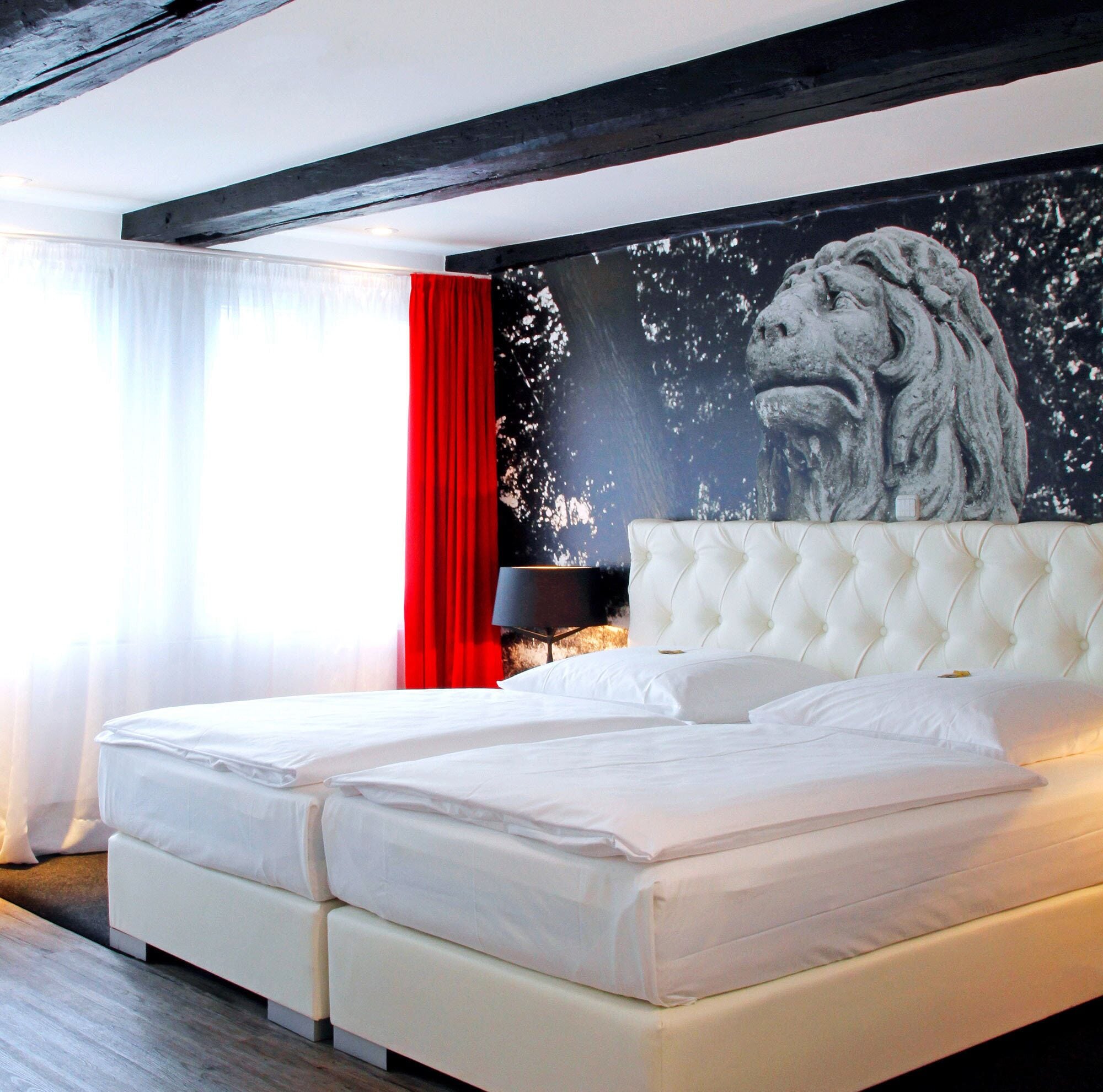 Ein Doppelbett mit weißen Leinen in einem modernen Zimmer, das durch ein großes Wandgemälde mit dem Motiv eines Löwenkopfes gekennzeichnet ist.