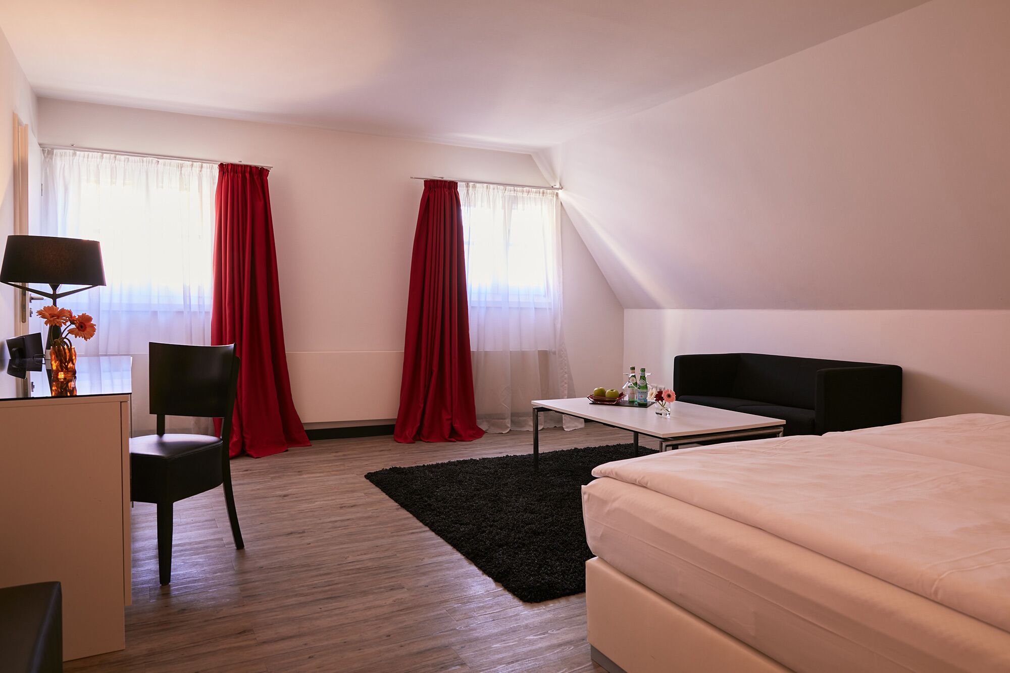 Una habitación de hotel moderna con una cama, un escritorio con lámpara, una silla, una mesita con un florero y dos ventanas con cortinas rojas.