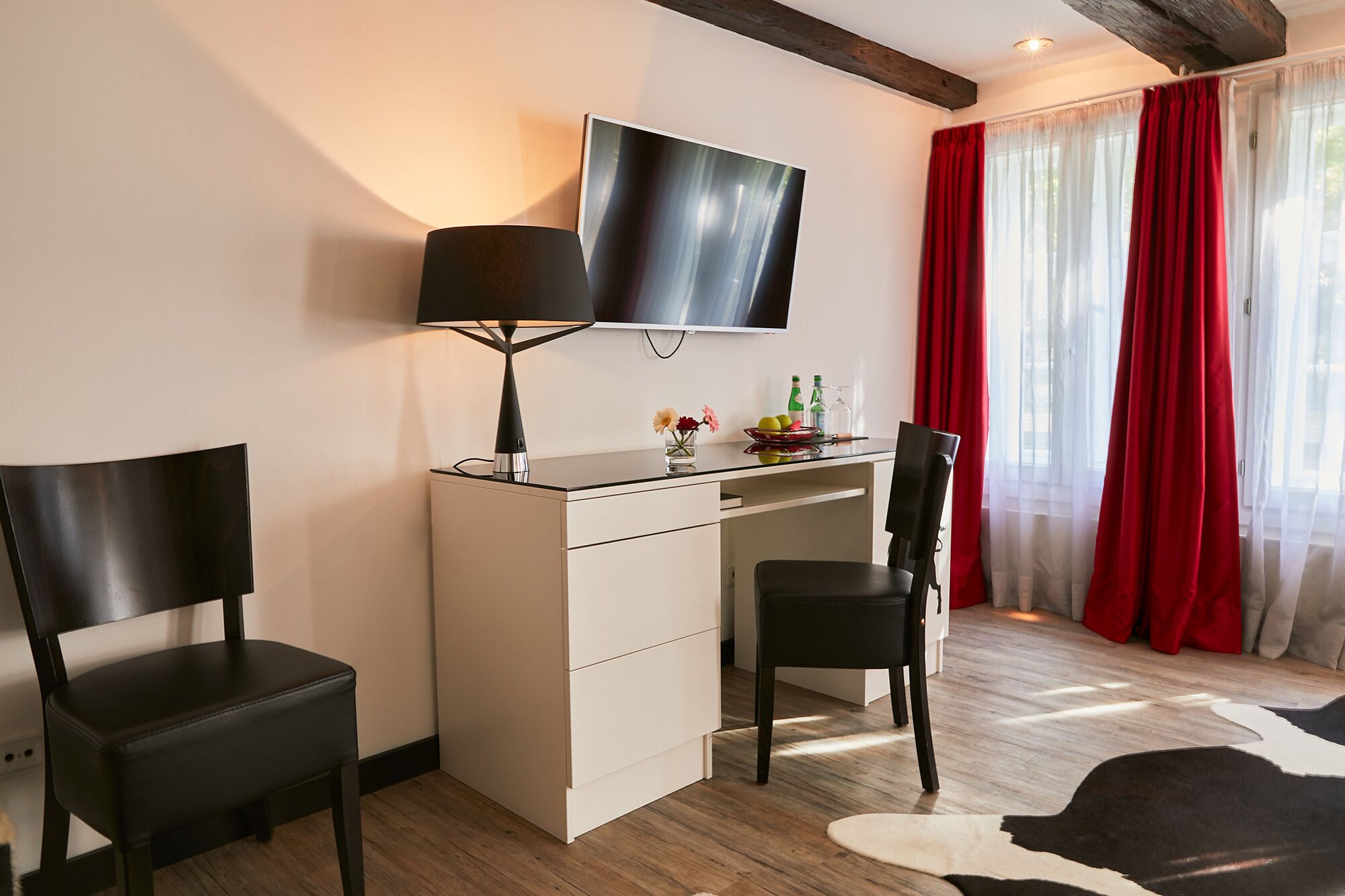 Una habitación moderna con escritorio, dos sillas, televisor y ventana con cortinas rojas.
