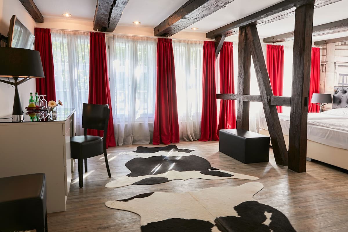 Ein modernes Hotelzimmer mit Holzboden, einem großen Fenster mit roten Vorhängen, einer schwarzen Ledersofa und einem weißen Bett sowie einem Kuhfellteppich.