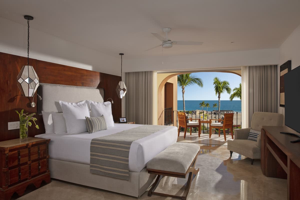 A bedroom with a large bed, a nightstand, a dresser, a TV, and a balcony with a view of the ocean.