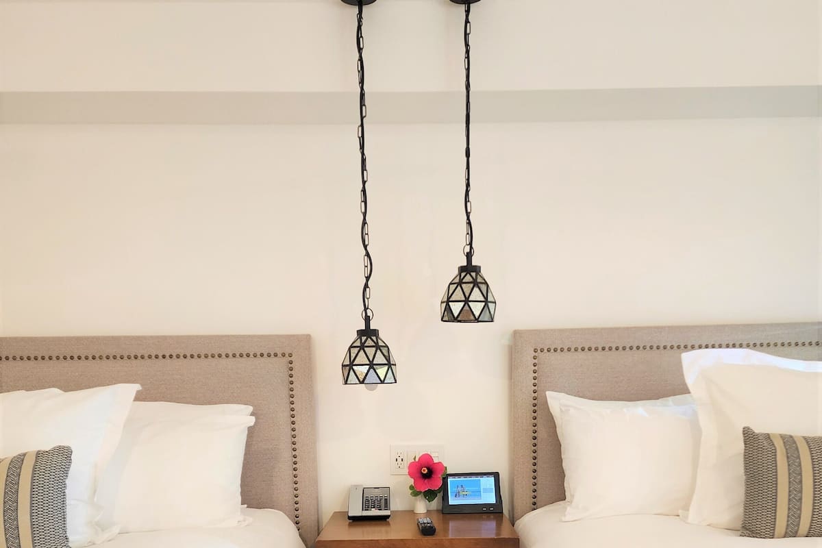 A hotel room with two beds, a nightstand with a phone and remote, and two hanging lamps.