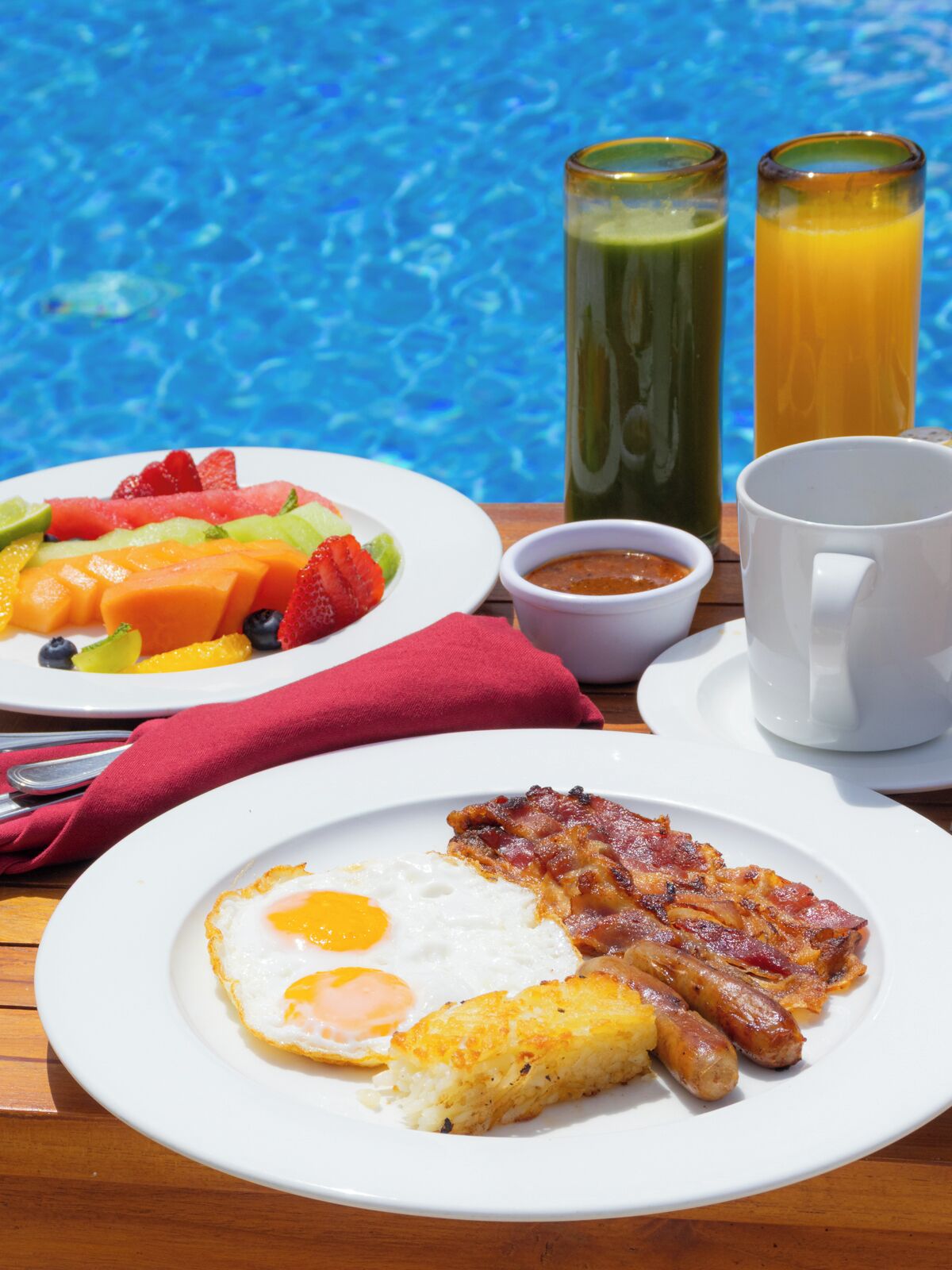 Un desayuno con un plato de huevos, tocino y salchichas, un plato de fruta y dos bebidas.
