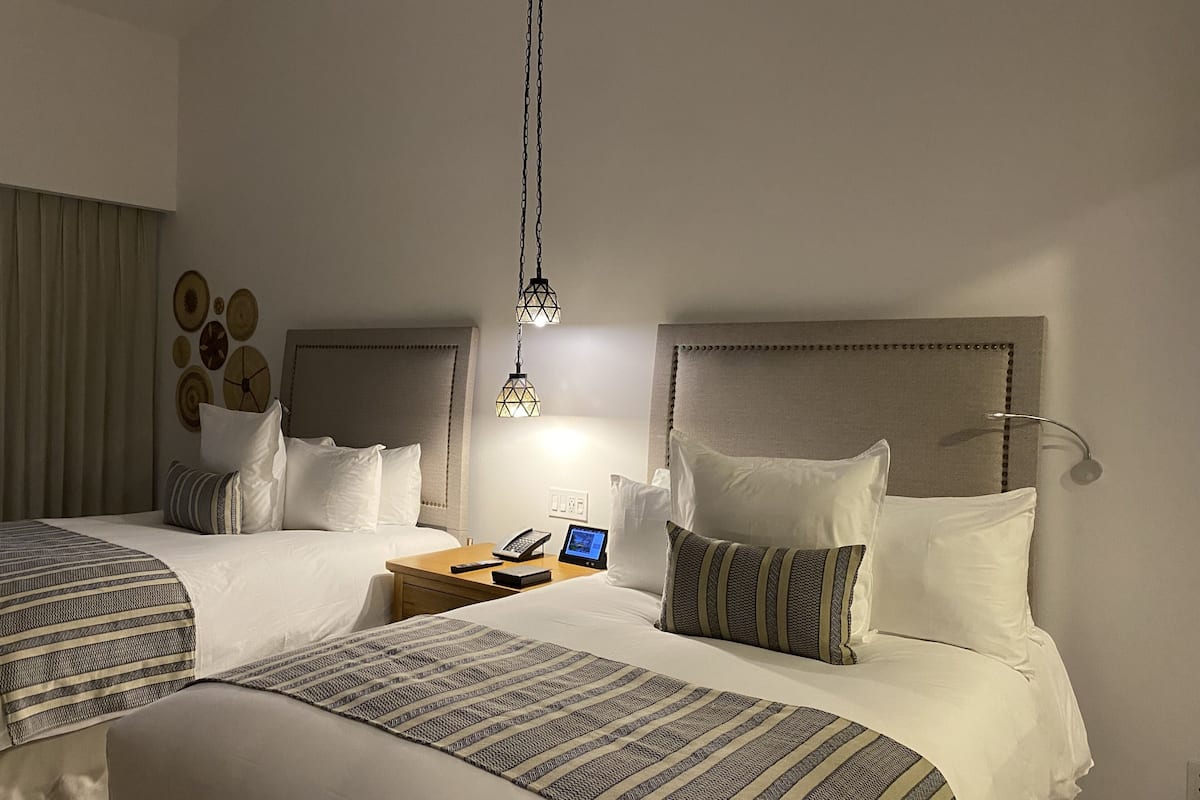 A hotel room with two beds, a bedside table with a phone and a tablet, and a wall-mounted lamp.
