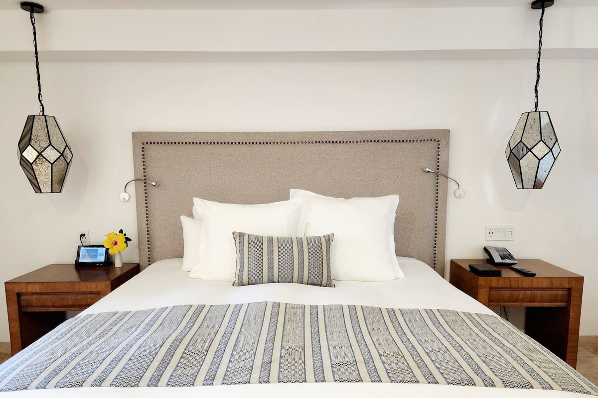 A neatly made bed with white bedding and a patterned throw, flanked by two nightstands with lamps, a phone, and a small table with a flower vase.