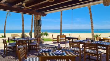 Un restaurant a la vora de la platja amb taules i cadires de fusta, un pilaster de pedra i vista a l'oceà.