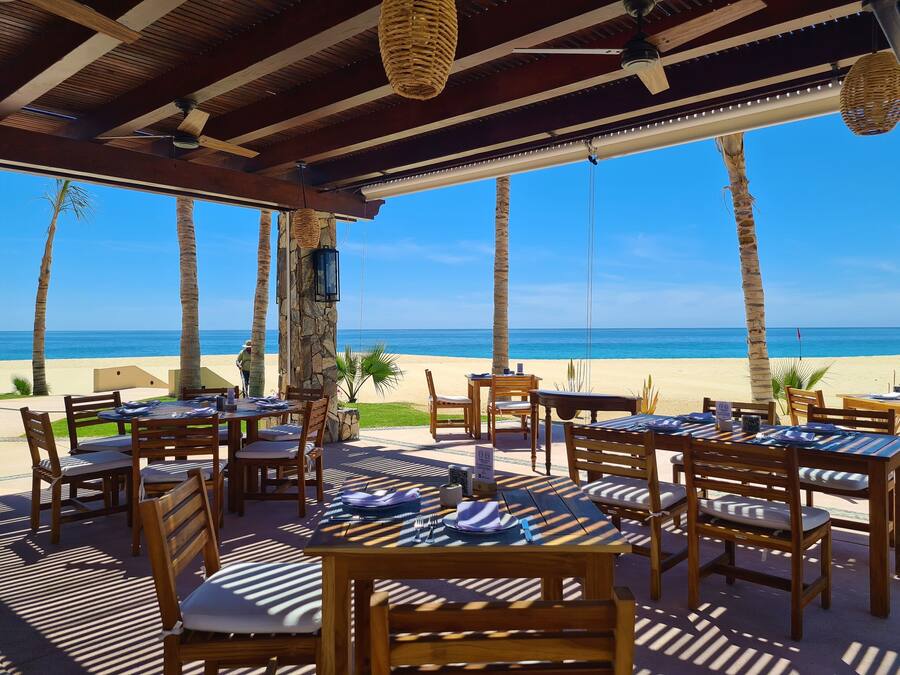 Un restaurant a la vora de la platja amb taules i cadires de fusta, un pilaster de pedra i vista a l'oceà.