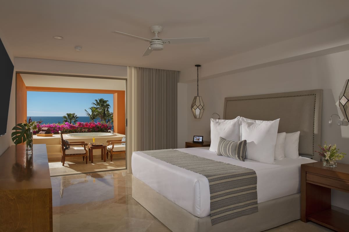 A modern hotel room with a large bed, a ceiling fan, a view of the ocean, a dining area with a table and chairs, and a small side table with a vase.
