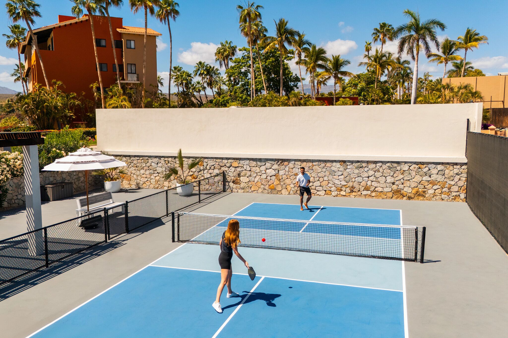 Un court de tennis en plein air, avec un mur en pierre et des palmiers en arrière-plan.
