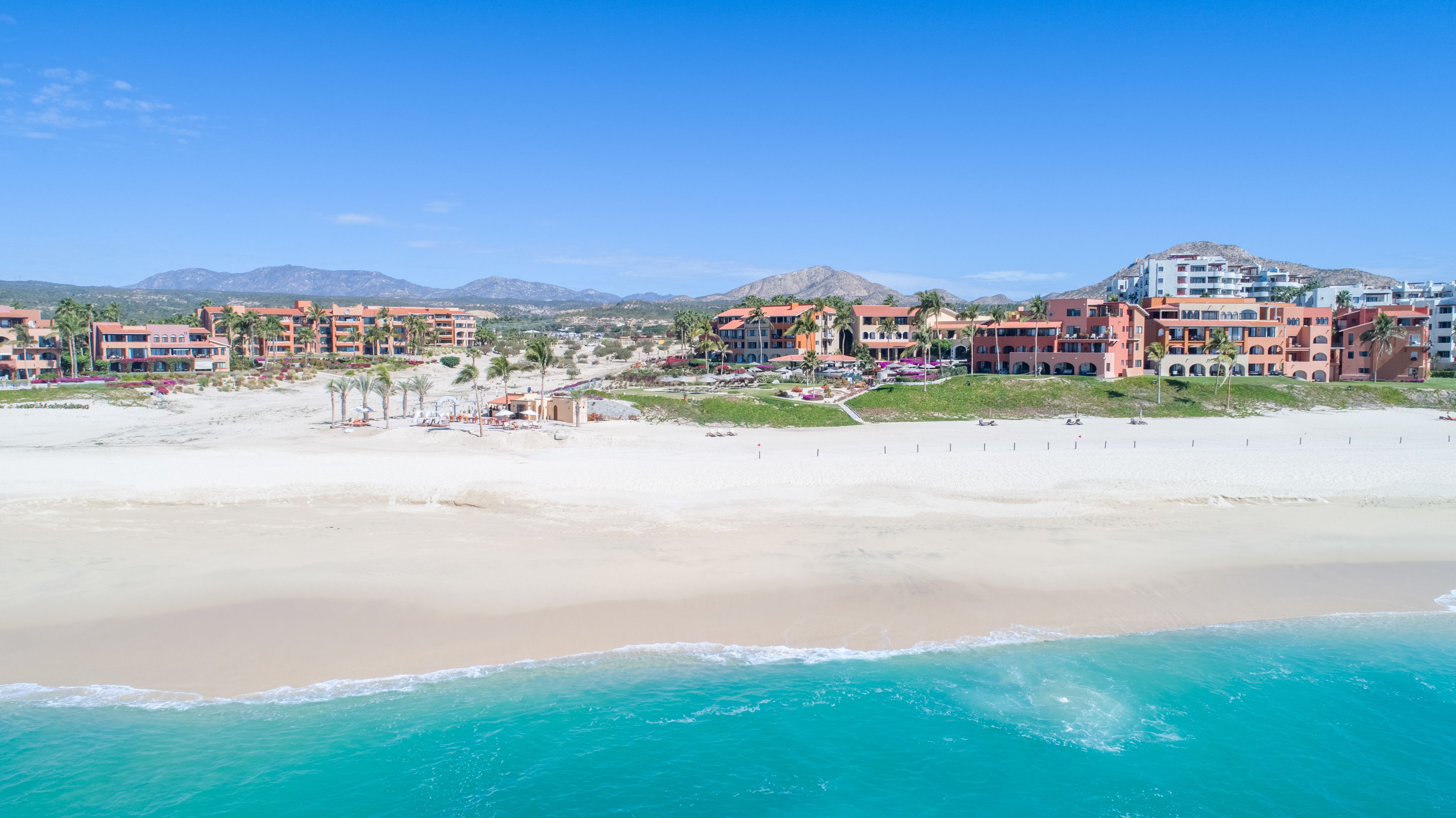 Un hotel frente a la playa con varias edificaciones, una playa de arena y un océano azul cristalino.