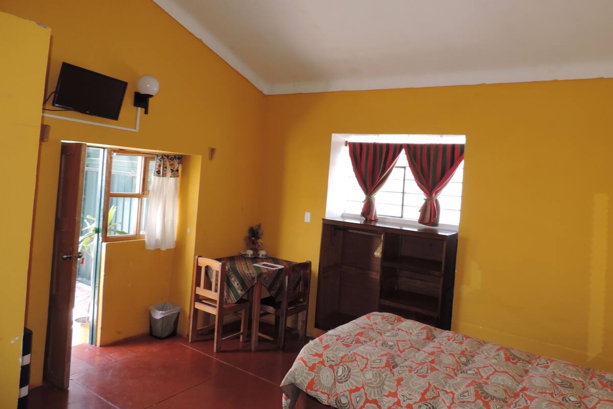 Ein Zimmer mit gelben Wänden, einem Bett mit gemustertem Überwurf, einem Holztisch mit Stuhl, einem kleinen Tisch mit einer Vase und einem Fenster mit Vorhängen.