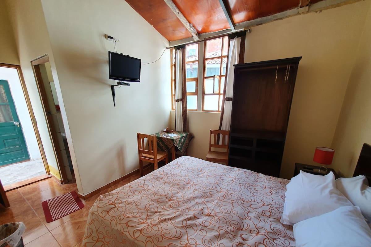 Ein Schlafzimmer mit einem Bett, einem an der Wand montierten Fernseher, einem Holzstuhl, einem kleinen Tisch und einem Fenster mit Vorhängen.