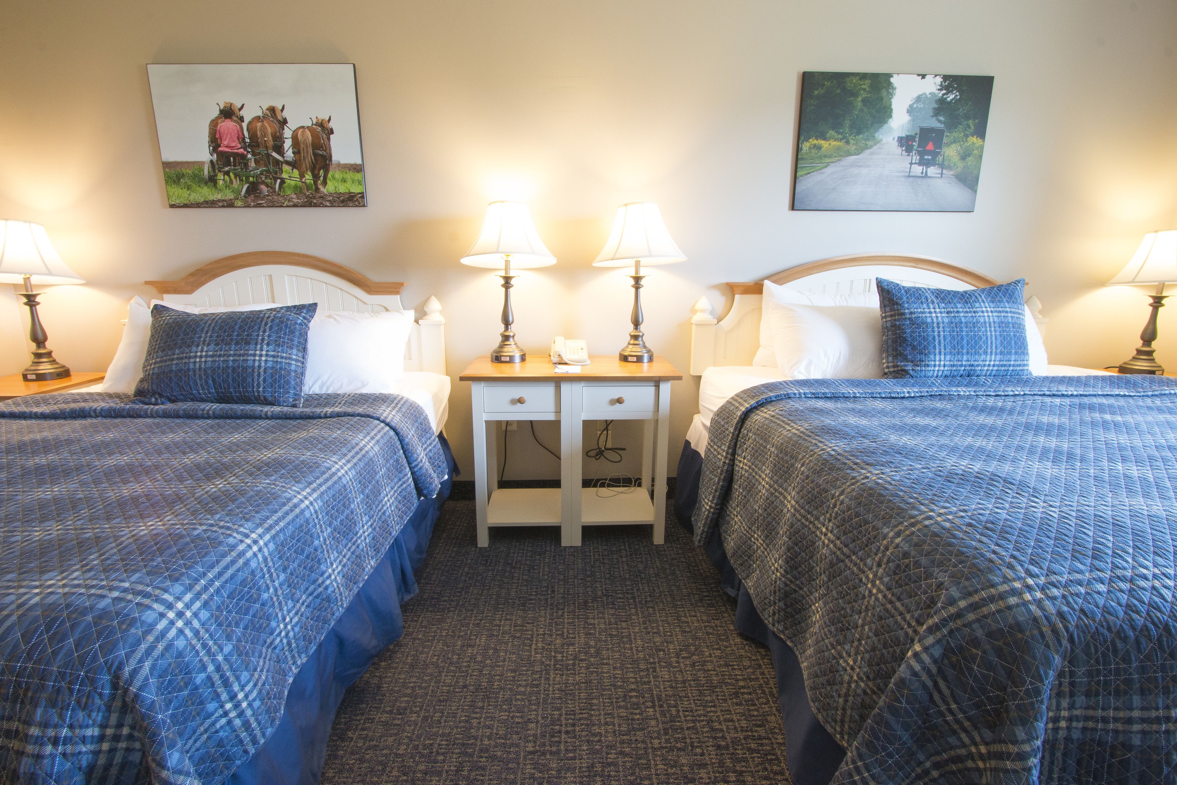 Habitación de hotel con dos camas, cada una con cubrecama de cuadros azules, una mesita de noche con dos lámparas y dos cuadros colgados en la pared.