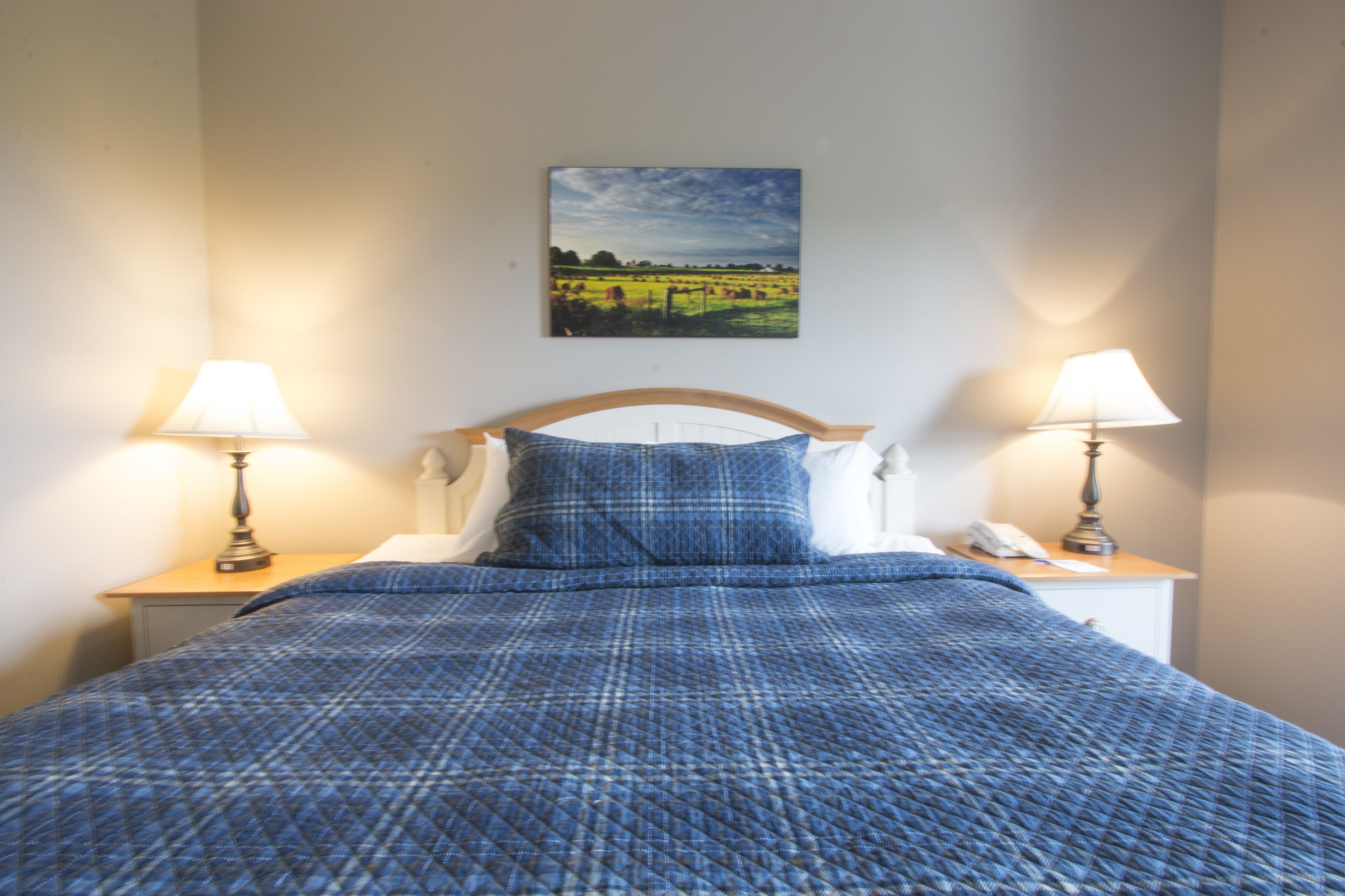 A neatly made bed with a blue quilt, two bedside lamps, a white telephone, and a landscape painting on the wall.