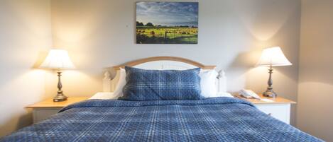A neatly made bed with a blue quilt, two bedside lamps, a white telephone, and a landscape painting on the wall.