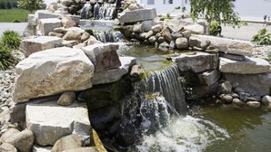A waterfall feature with large rocks and a small pond, surrounded by a landscaped area with a white building in the background.