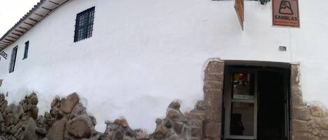 A white building with a stone wall at the base, a sign for 'SANBLAS Backpacker Hostel', and a window with green shutters.