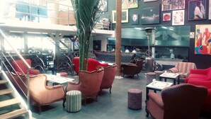 Un espacio interior amplio con diversas opciones de asiento, incluyendo sofás y sillones, dispuestos alrededor de una columna central con una planta. Se observa una escalera a la izquierda y vistas al exterior a través de amplias ventanas.