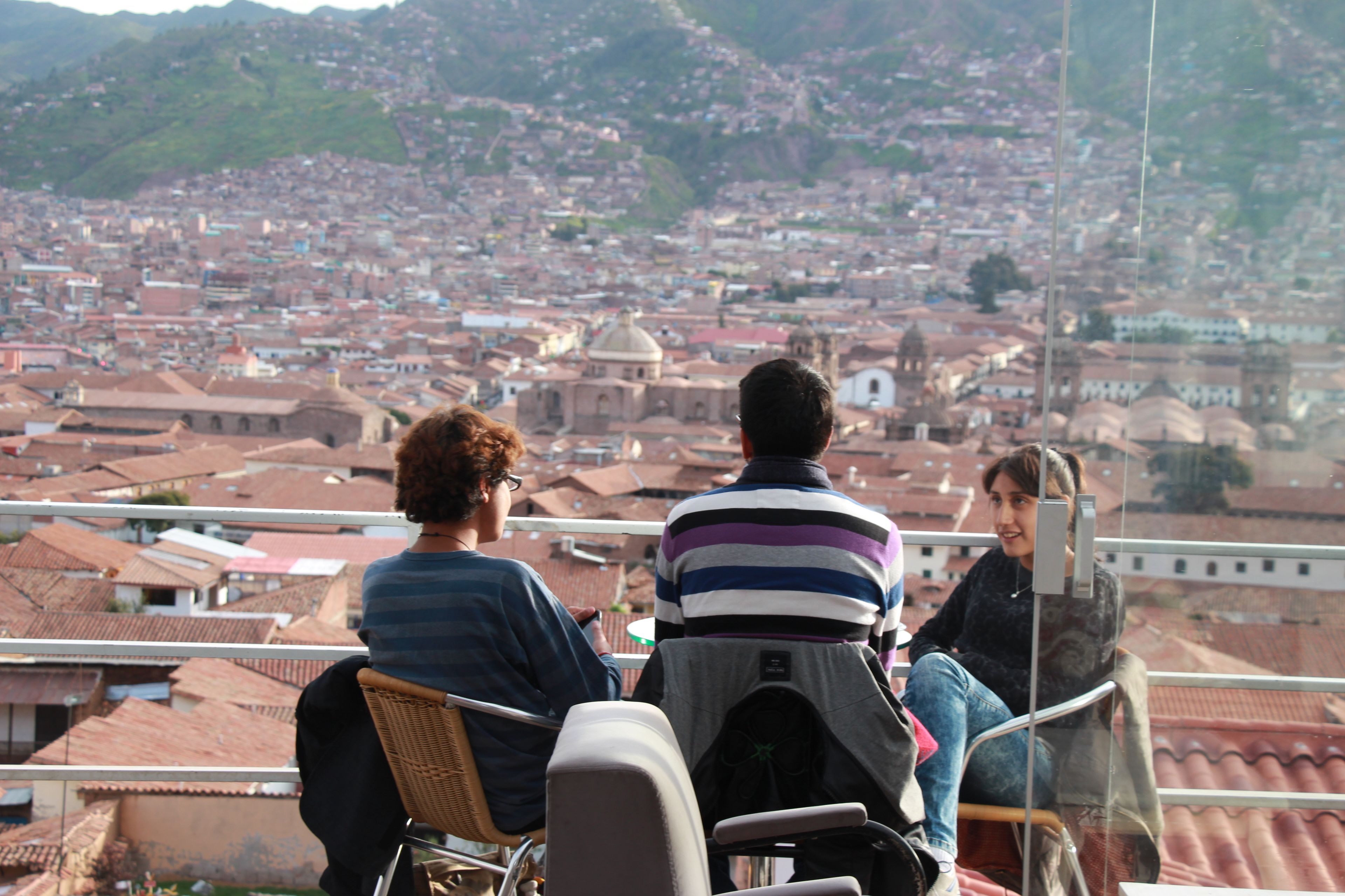 Trois personnes sont assises sur un balcon qui donne sur une vue panoramique de la ville, avec des bâtiments aux toits rouges.