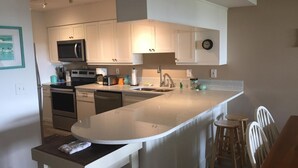 A modern kitchen with white cabinetry, a central island, and a dining area with wooden chairs. - Ocean Gate at Hibiscus Resort (St. Augustine)