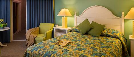 Habitación de hotel con una cama, dos mesitas de noche, una silla y una puerta que da a otra habitación.