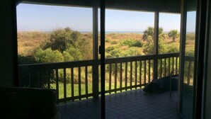 A room with a sliding glass door leading to an outdoor balcony with a view of a grassy landscape and trees. - Ocean Gate at Hibiscus Resort (St. Augustine)